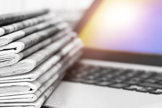 A stack of folded black-and-white newspapers lies on the keyboard of a laptop with an illuminated screen in the background