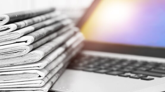 A stack of folded black-and-white newspapers lies on the keyboard of a laptop with an illuminated screen in the background