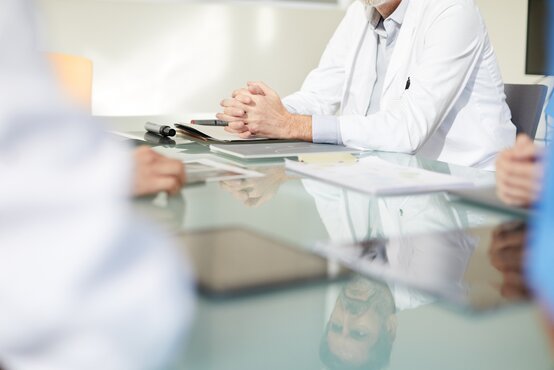 Several people in white coats are sitting at a glass table with documents and tablets, focus on hands clasped in the front