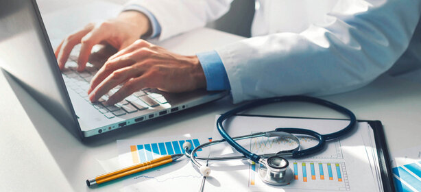 Hands of a person in a white coat typing on a laptop keyboard, stethoscope, pen and medical charts on a desk