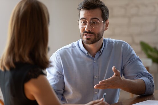 Junger Mann mit Bart und Brille spricht im modernen Büro mit einer Frau, beide am Tisch, helle Wand und Pflanze im Hintergrund
