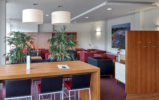 Spacious, modern patient lounge with large wooden table, black and red chairs, pendant lamps, red flower arrangements and armchairs