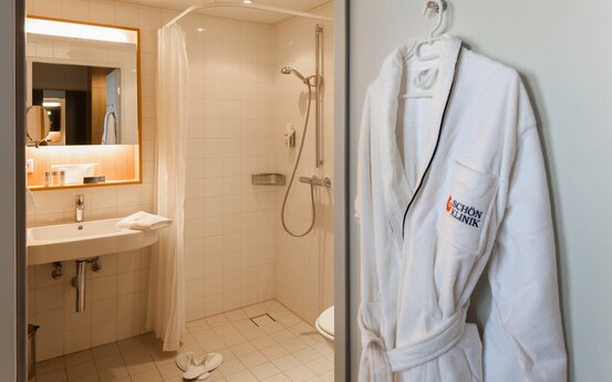 Modern clinic bathroom with large pale tiles, wall-mounted white sink, back-lit mirror, walk-in shower with white curtain, toiletries and towels