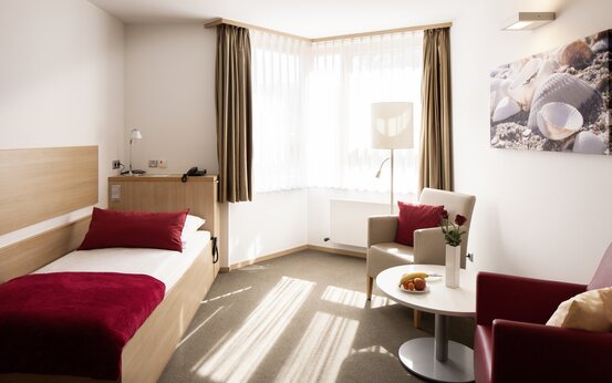 Bright, modern single patient room with wood furniture, large windows with sheer curtains, two lounge chairs and round table