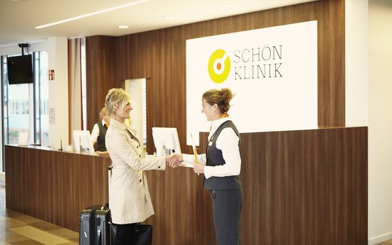 A patient with a wheeled suitcase is greeted warmly by a member of staff at the reception desk of the Schoen Clinic