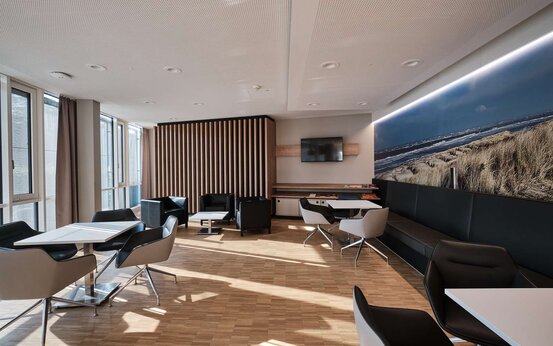 Heller Aufenthaltsraum mit Stühlen, Sesseln und Sitzbank. Fernseher und Bild von einer Küste im Hintergrund