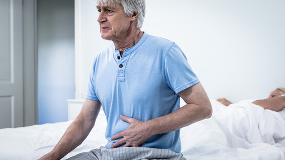 An elderly man sits on the edge of the bed with a pained expression on his face, holding his hand on his upper abdomen