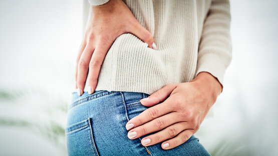 Close-up of a person wearing blue jeans and a cream-coloured jumper, holding both hands on their lower back and hips