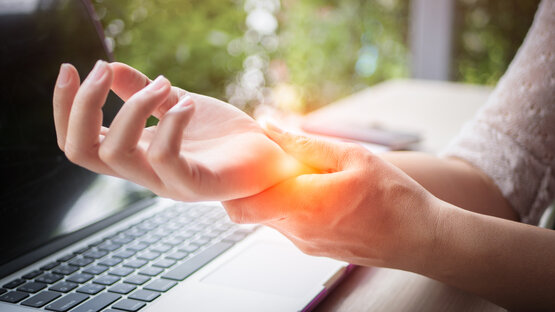 Person holding painful right wrist with left hand above laptop keyboard, orange light reflection indicates pain