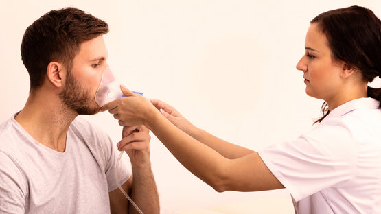 Pflegekraft hält einem sitzenden Mann eine Sauerstoffmaske vor das Gesicht zur Inhalationsbehandlung im hellen Behandlungsraum