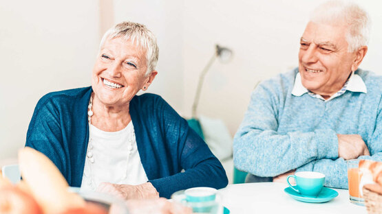 Two elderly people are sitting at a table with blue cups and juice glasses, both laughing.