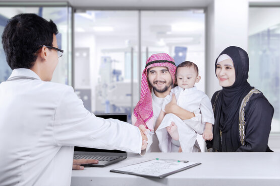 Doctor in white coat shakes hands with father in traditional robe and headscarf, mother with child standing next to them