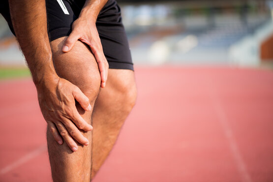 Man in black sports shorts grabs his sore knee with both hands on a red running track, indicating acute knee pain during exercise