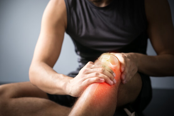 Man in black sleeveless sportswear sits upright indoors, grasping his right knee with both hands, red glow and anatomical overlay highlight knee