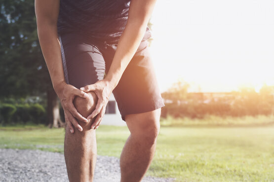 Man wearing athletic shorts and shirt outdoors, bending over and clutching his knee in pain on a gravel path in a sunny park
