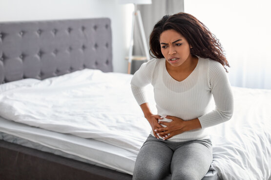 Young woman sitting on bed, holding her right upper abdomen in pain