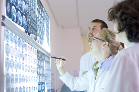 Three doctors in white coats look together at MRI images of a brain on an illuminated wall, person points to the images with a pen