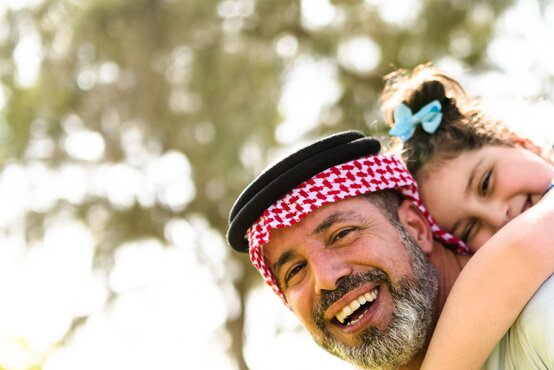 Smiling Arab father wearing a red and white kufiya, holding a child with a blue bow on his back