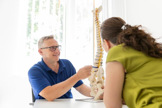 A doctor wearing glasses and a blue polo shirt explains anatomy to a patient using a model of the spine in a bright consultation room