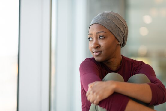 Woman in a grey headscarf and burgundy shirt sits by a window in a modern hospital, arms wrapped around her knees, looking thoughtfully outside