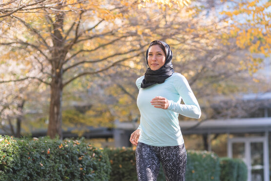 Woman in a black hijab, blue long-sleeved shirt, and patterned leggings jogs in a sunny autumn park