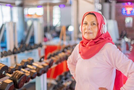 Older woman in a red hijab, pink workout top, and headphones stands smiling with hands on hips next to dumbbells in a modern gym