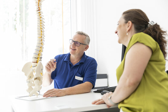 Doctor in blue polo shirt explains findings to patient using spinal model at desk