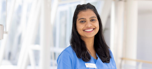 Medical staff member with long dark hair in a blue hospital uniform smiles at the camera in a modern, bright hospital corridor
