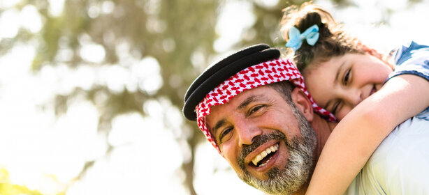 Smiling Arab father wearing a red and white kufiya, holding a child with a blue bow on his back